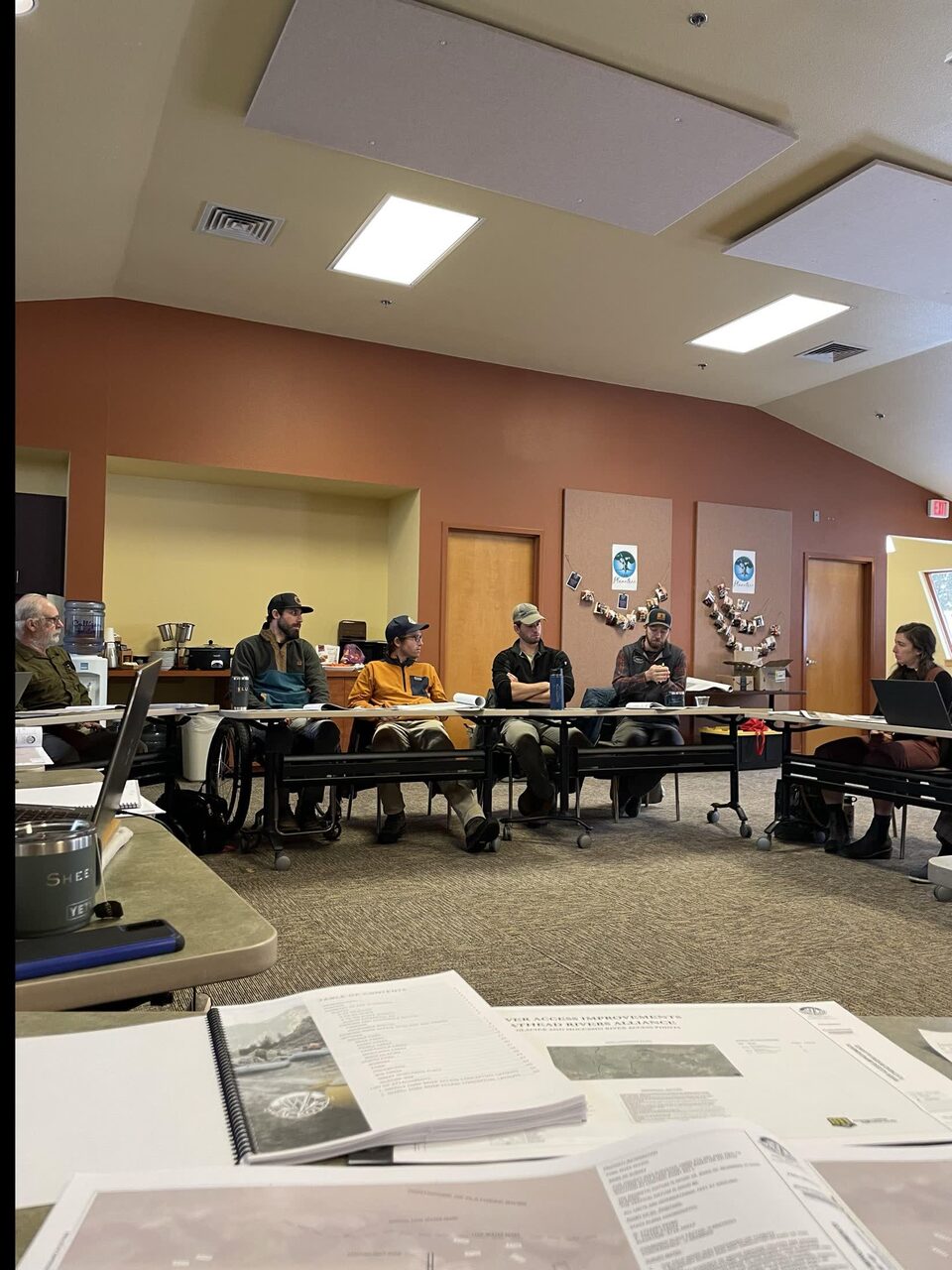 A planning meeting around long tables in a community room, a wheelchair pulled up to the table alongside the other seats, with printed Flathead river access reports open in the foreground.