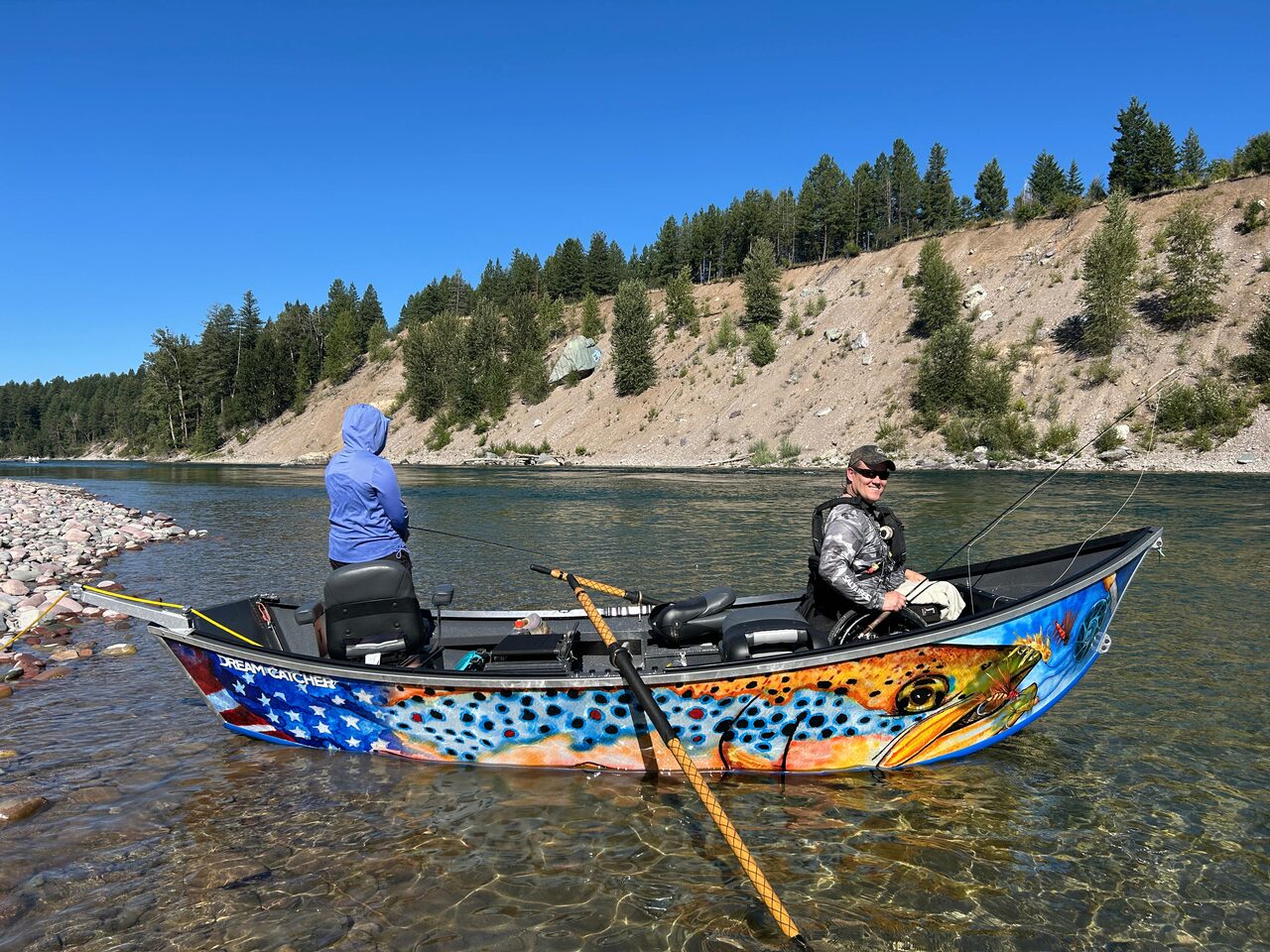 Two people in a drift boat on clear water, one standing in the bow and one at the oars, with a cobble bar in the foreground and a pale cut bank rising behind them.