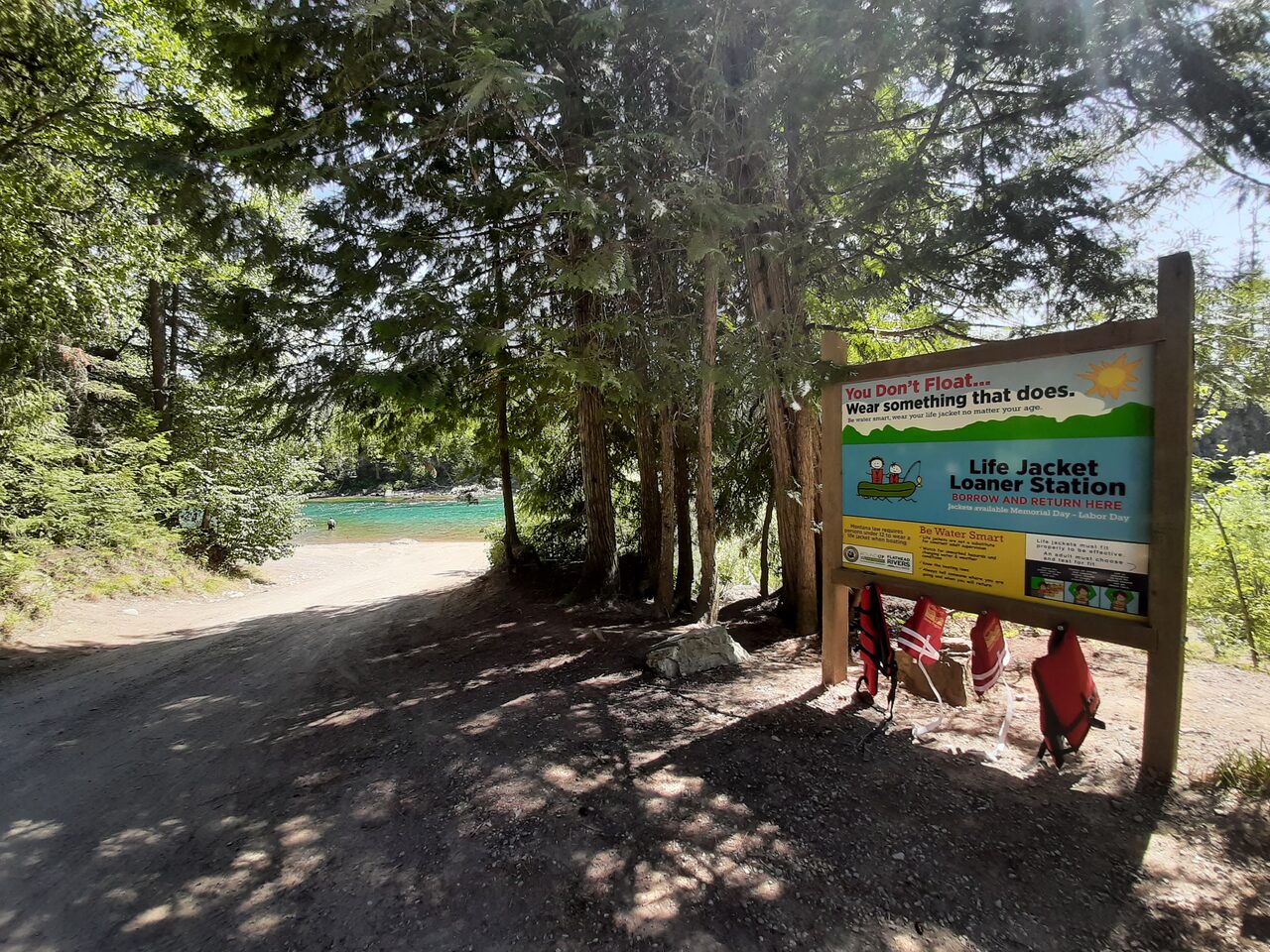 A life jacket loaner station at the head of a gravel access road: a timber kiosk hung with red life jackets under a sign reading Borrow and Return Here, with turquoise water visible through the cedars below.